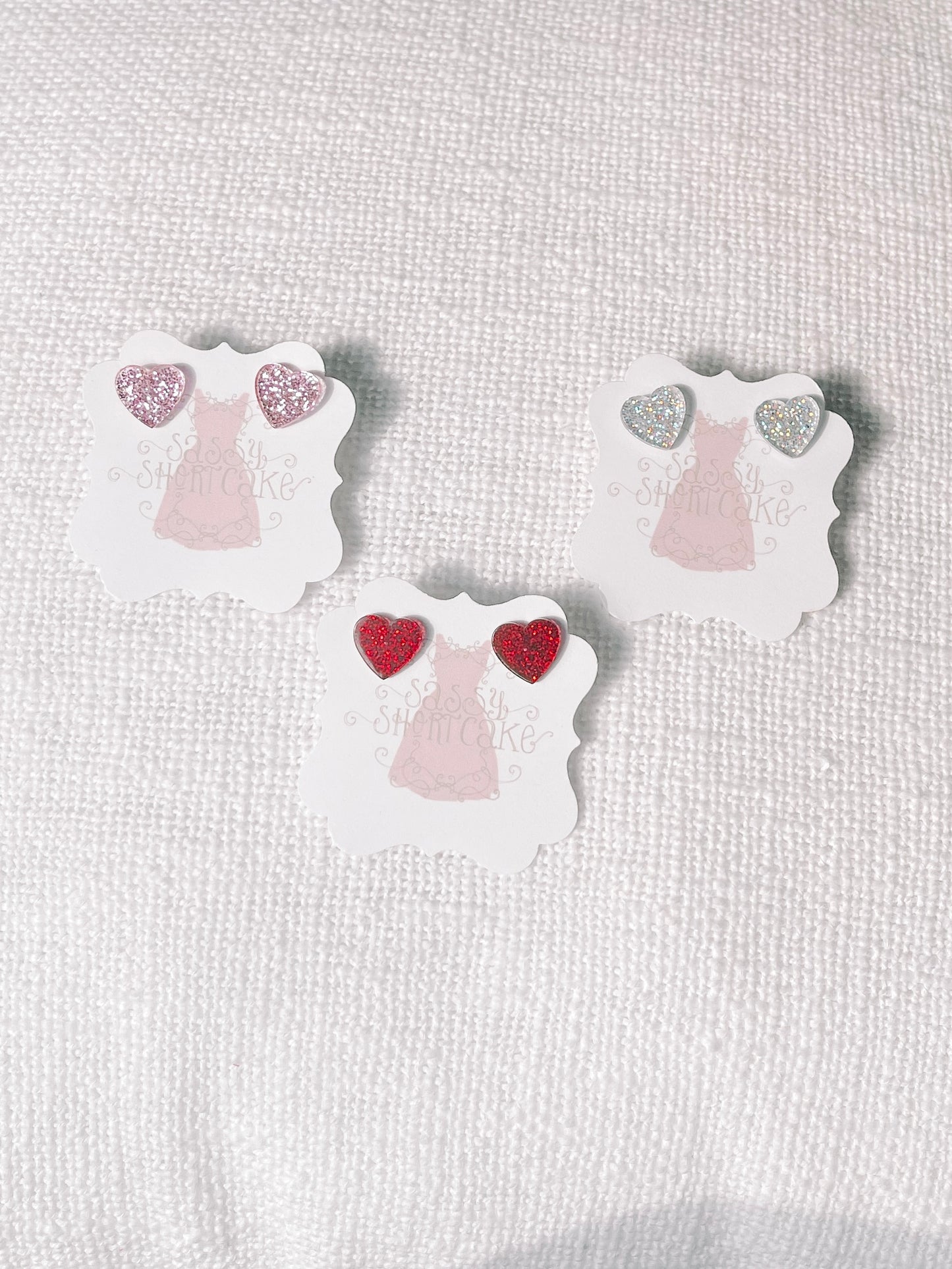 Glittery heart-shaped earrings displayed on individual cards, pink, silver, and red.