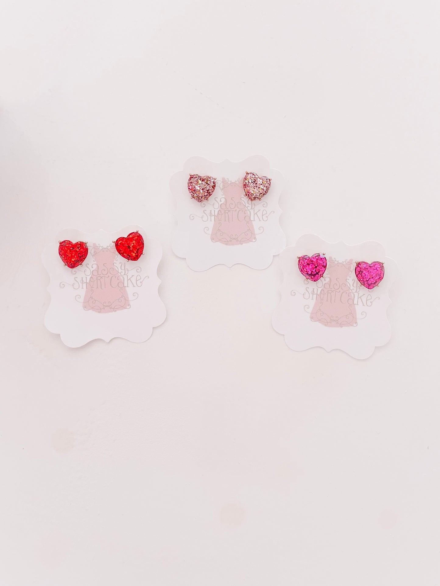 Sparkly heart-shaped earrings in red, pink, and rose gold displayed on individual cards.