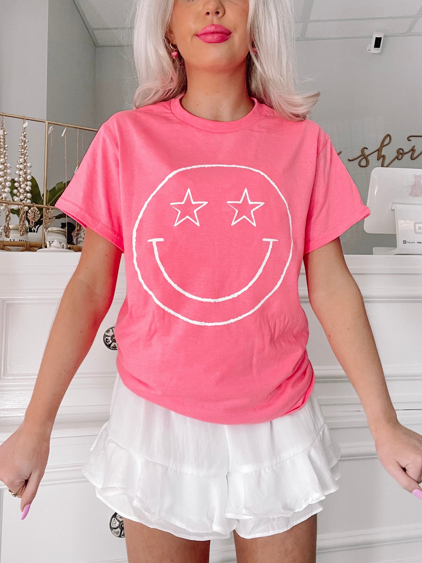 Pink short-sleeve tee featuring a white smiley face graphic; model is also wearing white ruffle shorts.