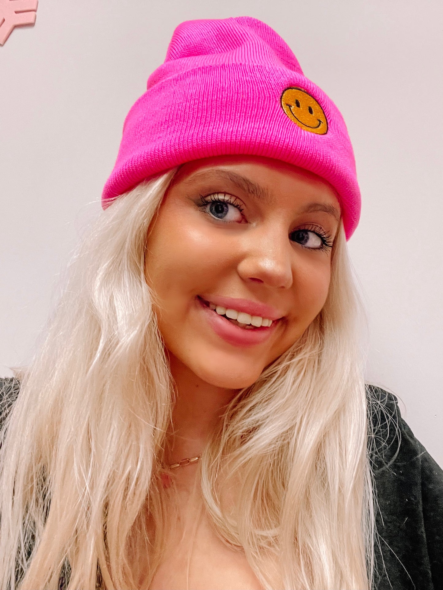 Hot pink beanie with a yellow smiley face, worn by a blonde woman.