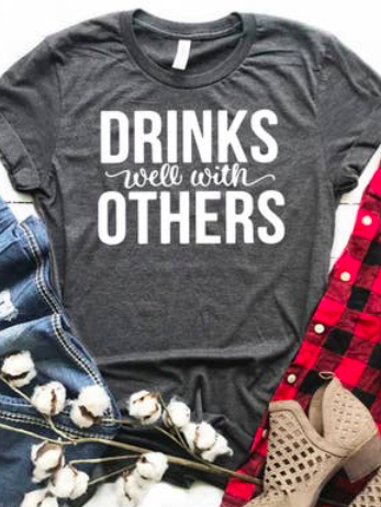 Dark gray tee shirt featuring the phrase "Drinks well with others" in white lettering, styled with denim and boots.