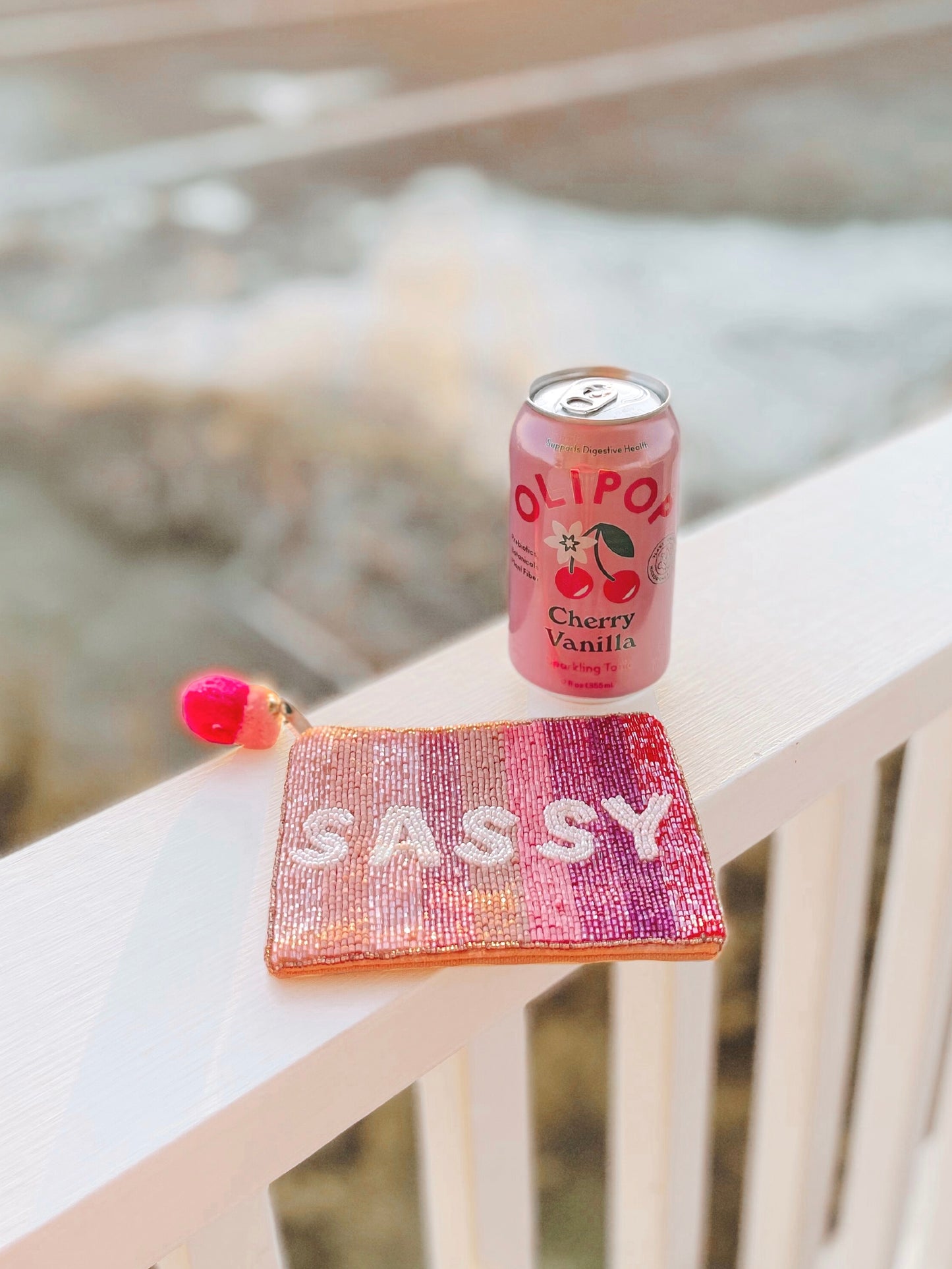 Beaded pouch with "SASSY" spelled out in white beads, pink and purple ombre design, resting on a white railing outdoors.