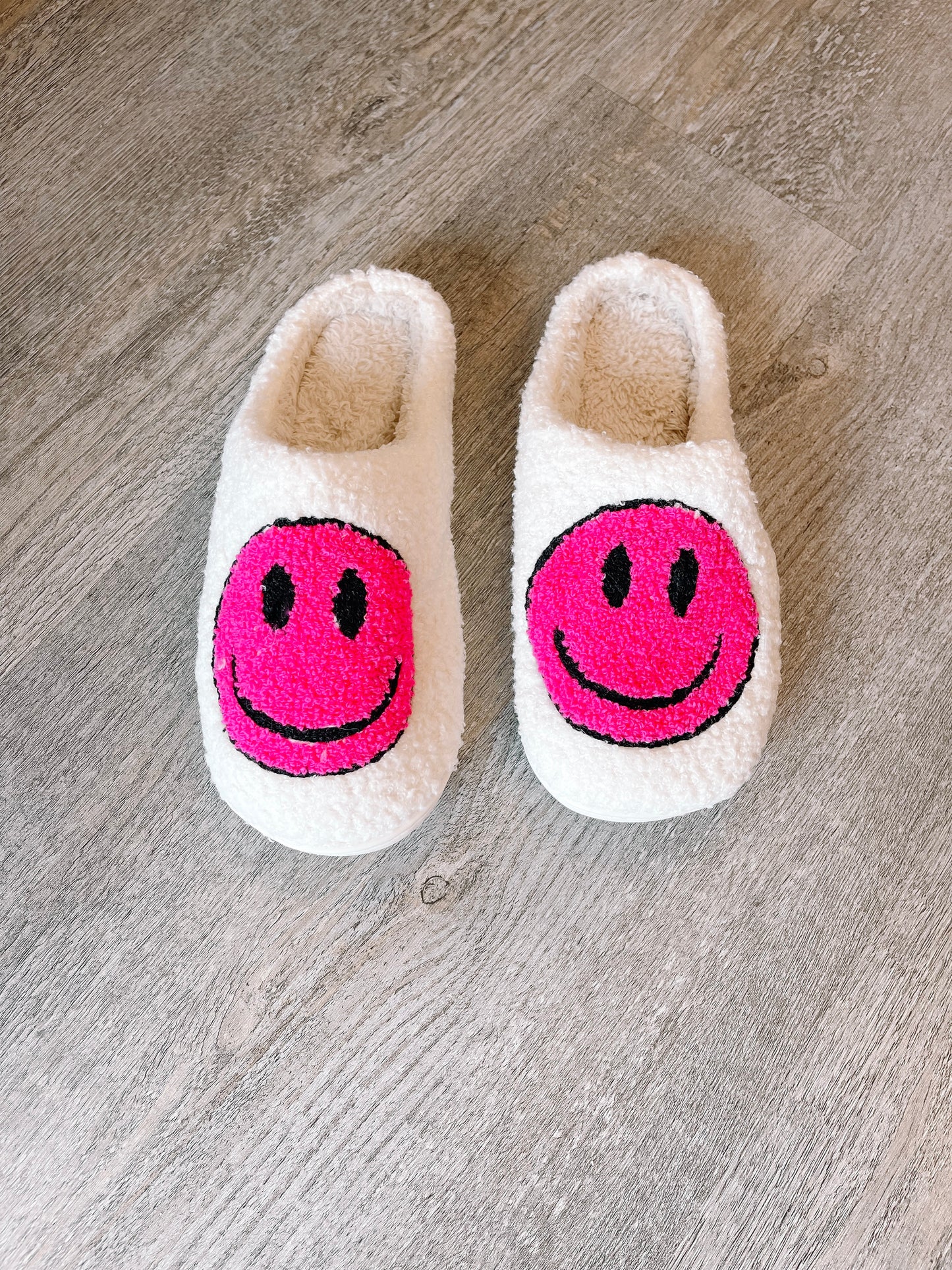 Fluffy white slippers with bright pink smiley faces, shown on a wood floor.