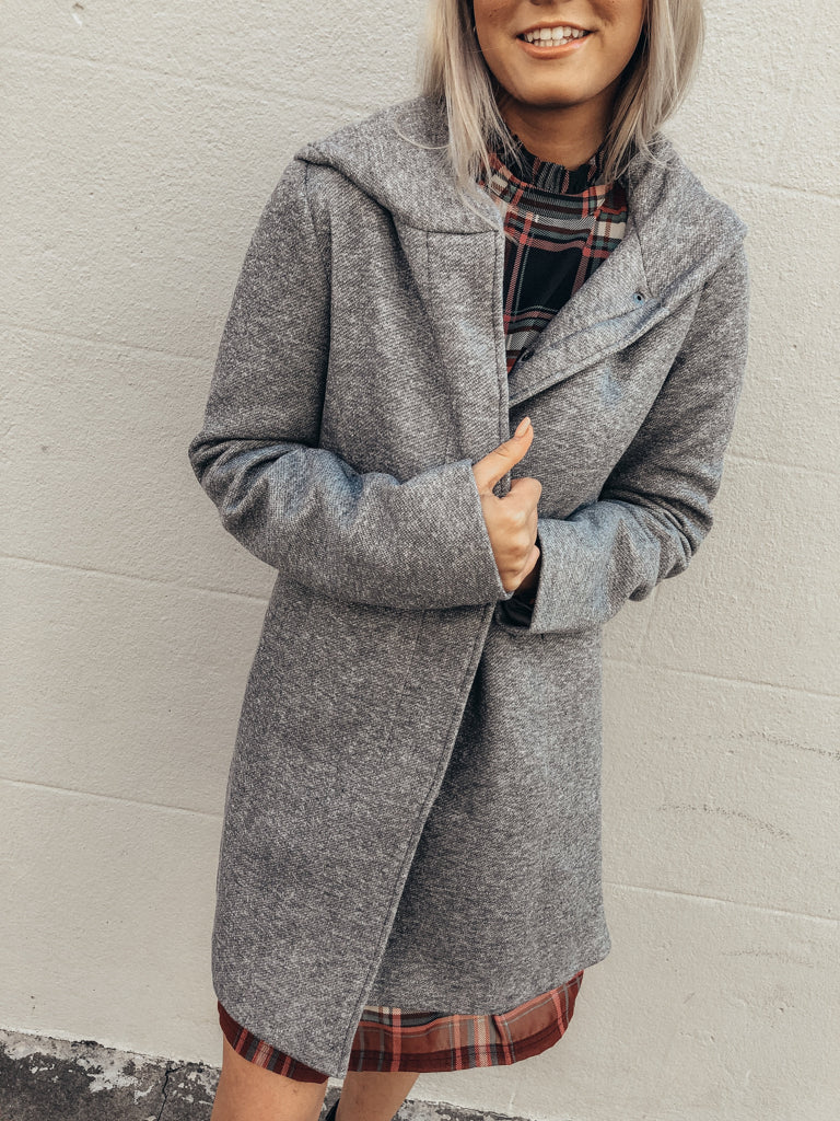 Gray hooded coat, heather texture, worn over a plaid dress against a white wall.