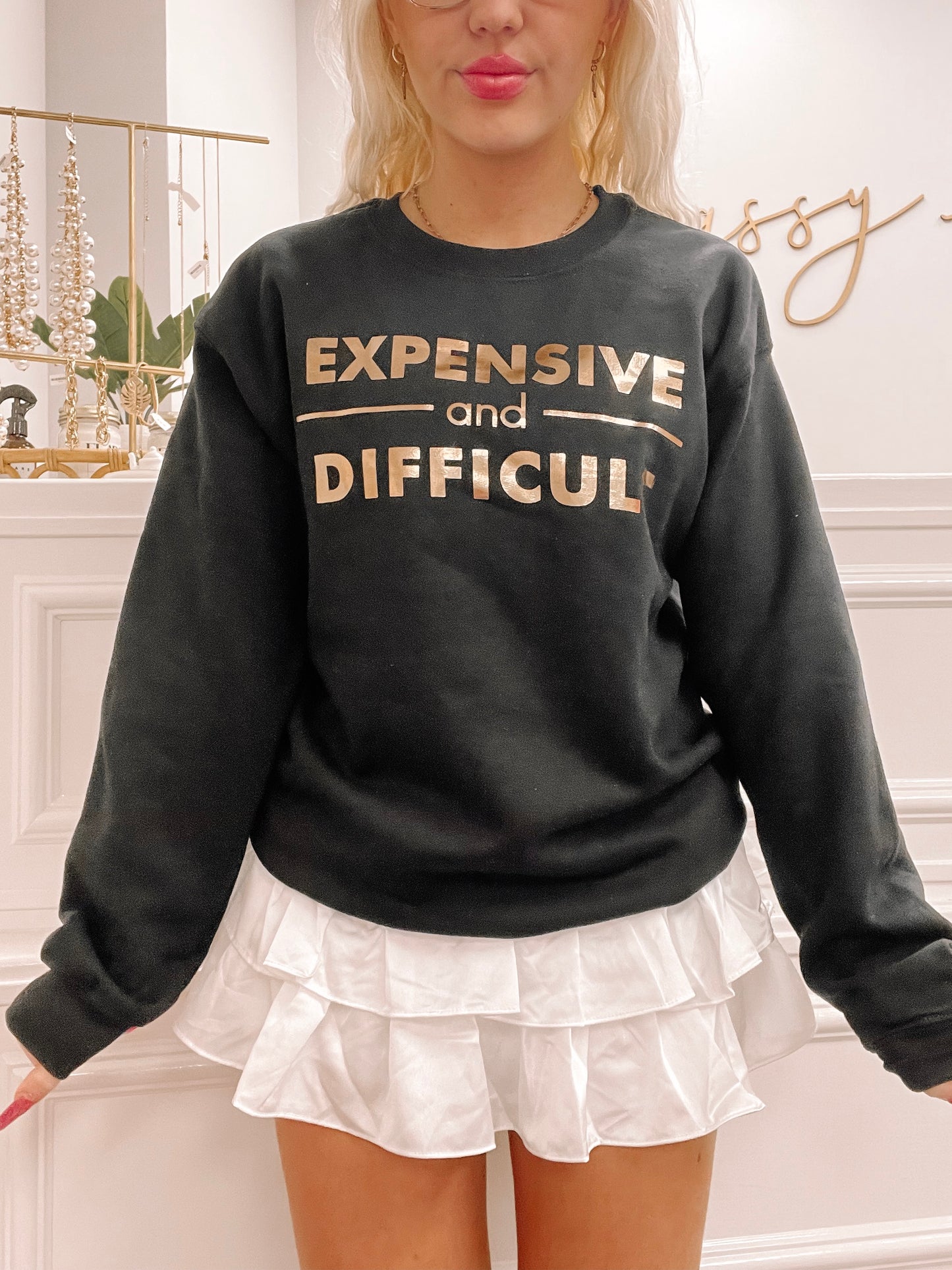 Black crewneck sweatshirt with rose gold "Expensive and Difficult" text; styled with white ruffled skirt.