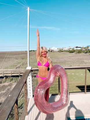 Woman in pink bikini on deck, holding glittery heart-shaped pool float; sunny outdoor scene.