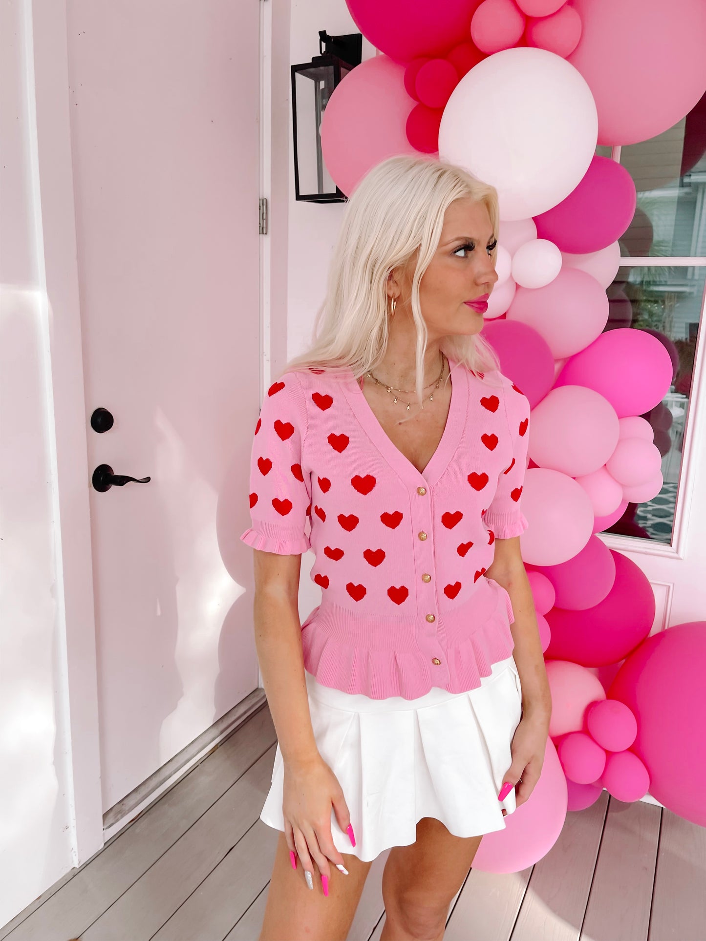 Pink button-up top with red heart pattern, modeled outdoors near pink balloon arch.