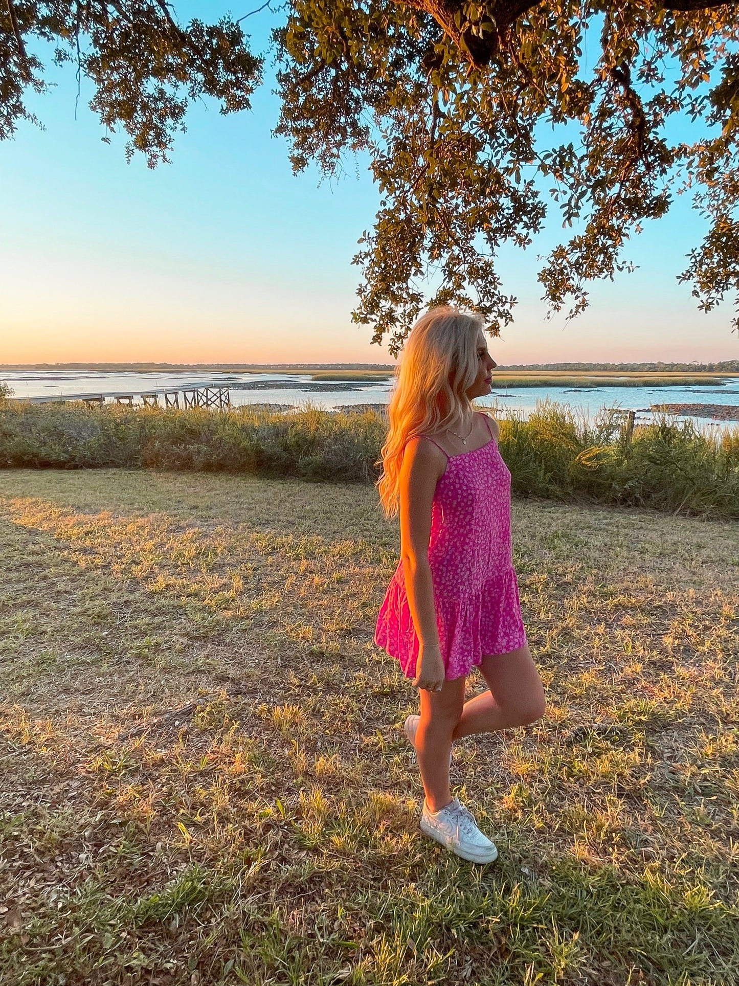 Woman in pink floral dress at sunset by the water.