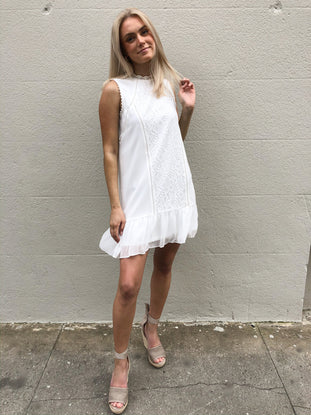 White eyelet dress with ruffle hem, styled with neutral wedges against a textured wall.