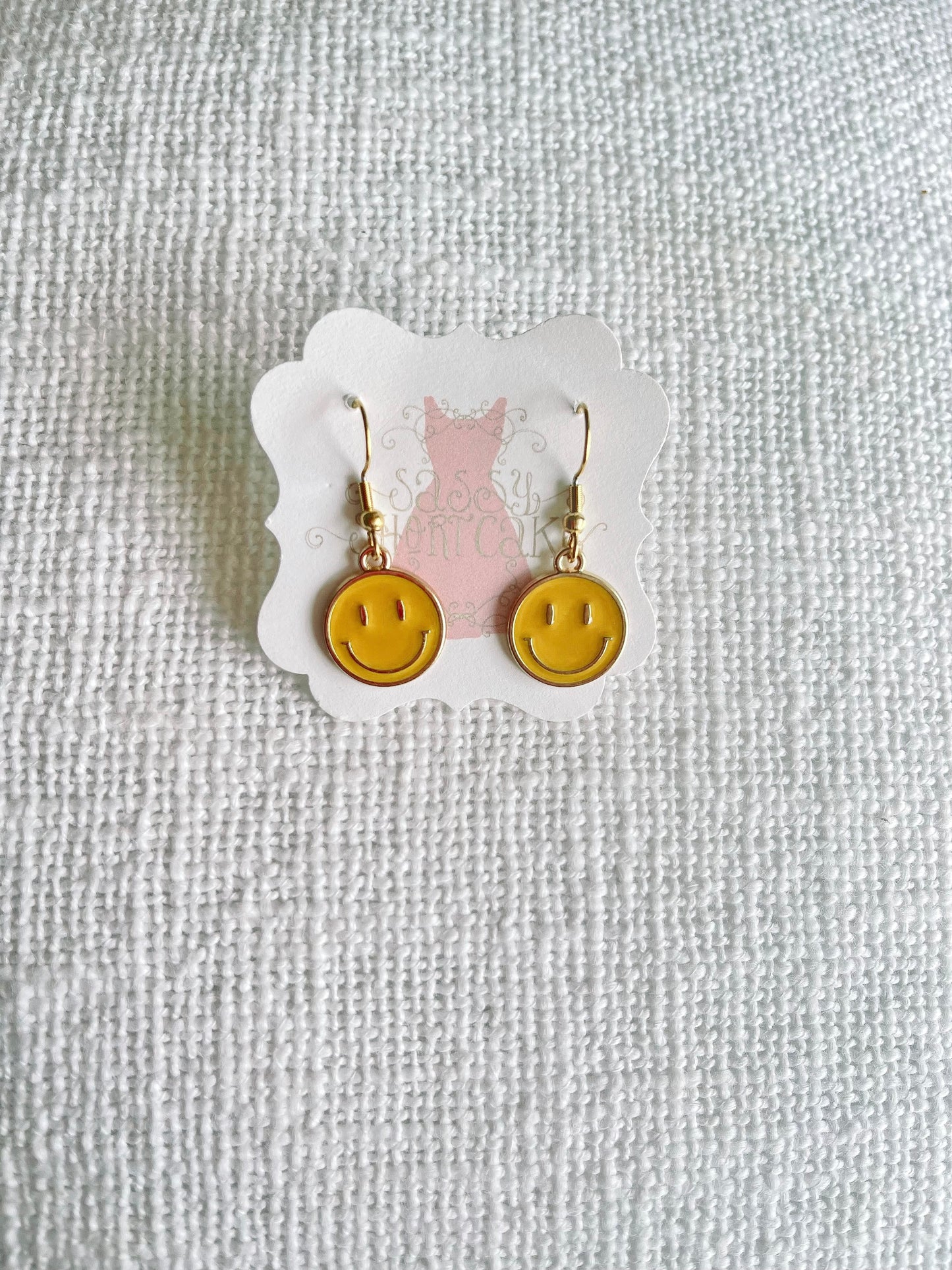 Gold smiley face dangle earrings on a white textured background.