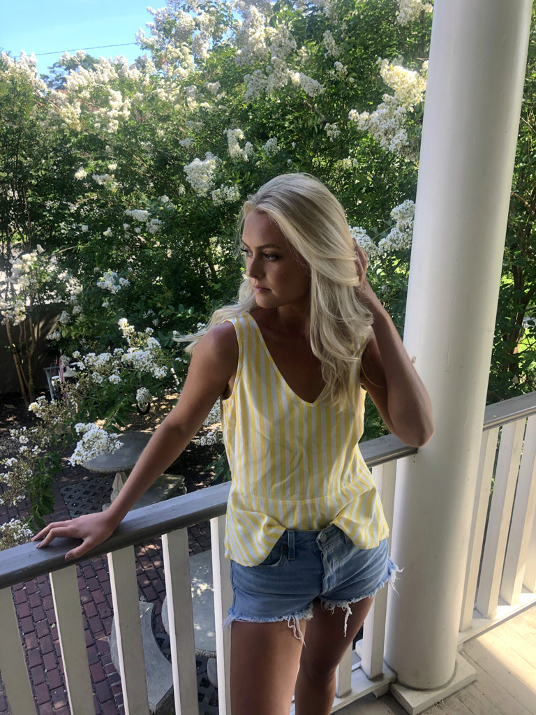 Woman in yellow striped tank top and denim shorts on a porch, blooming trees in the background.