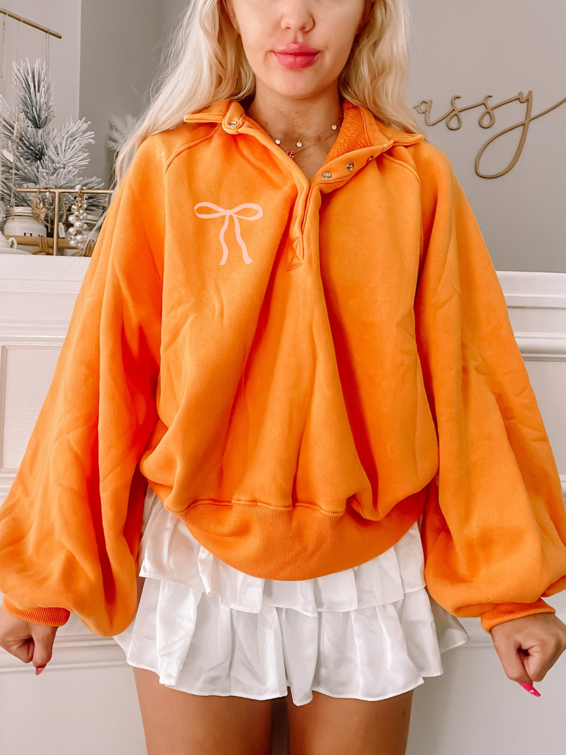 Orange pullover sweatshirt with a small pink bow detail, modeled with a white ruffled skirt.
