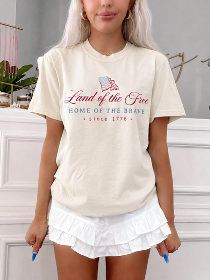 Land of the Free Tee from Sassy Shortcake in cream with patriotic front graphic, styled with white ruffle shorts on a blonde model.