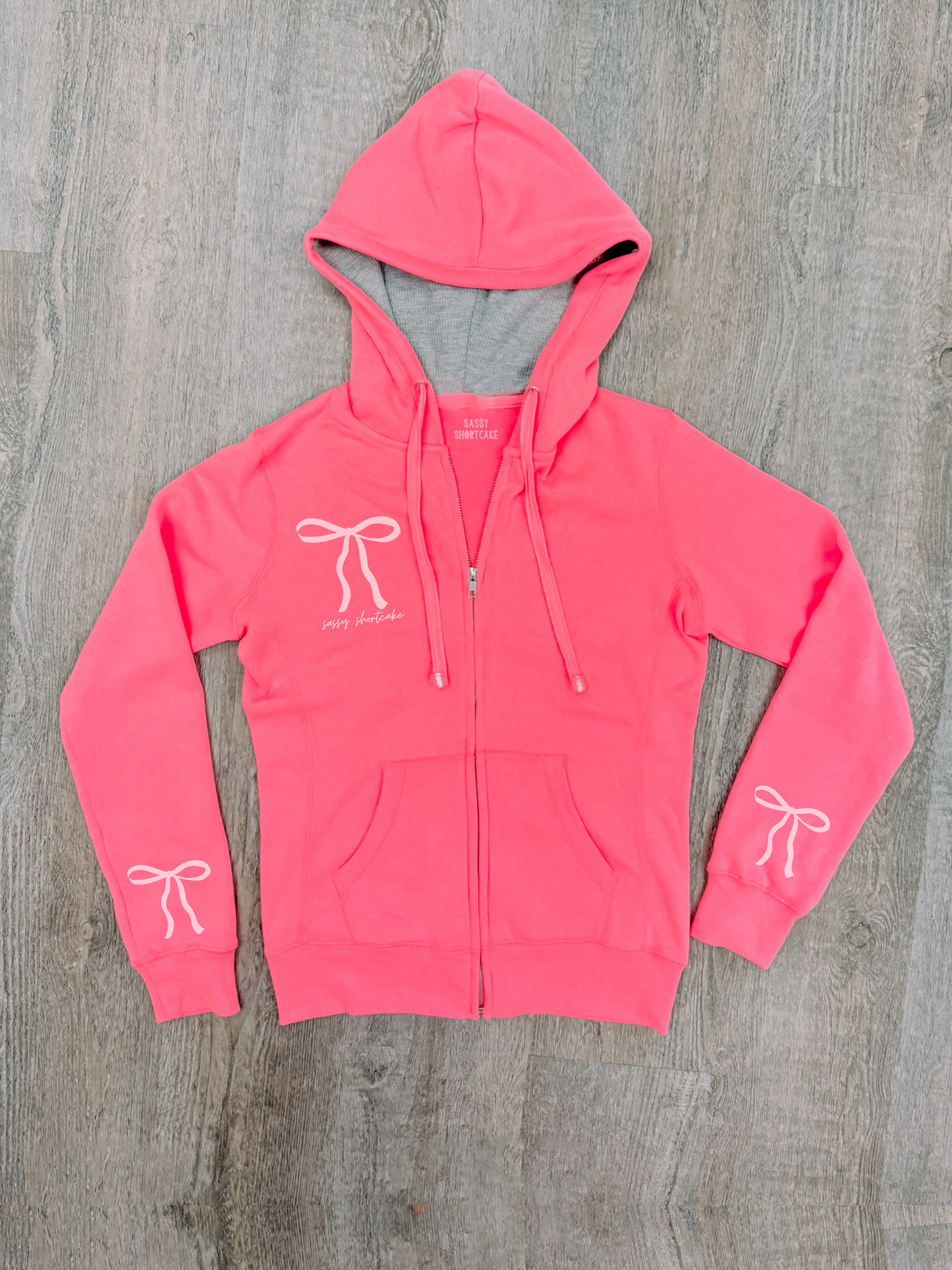 Neon pink zip-up hoodie with bow details, lying flat on wood.
