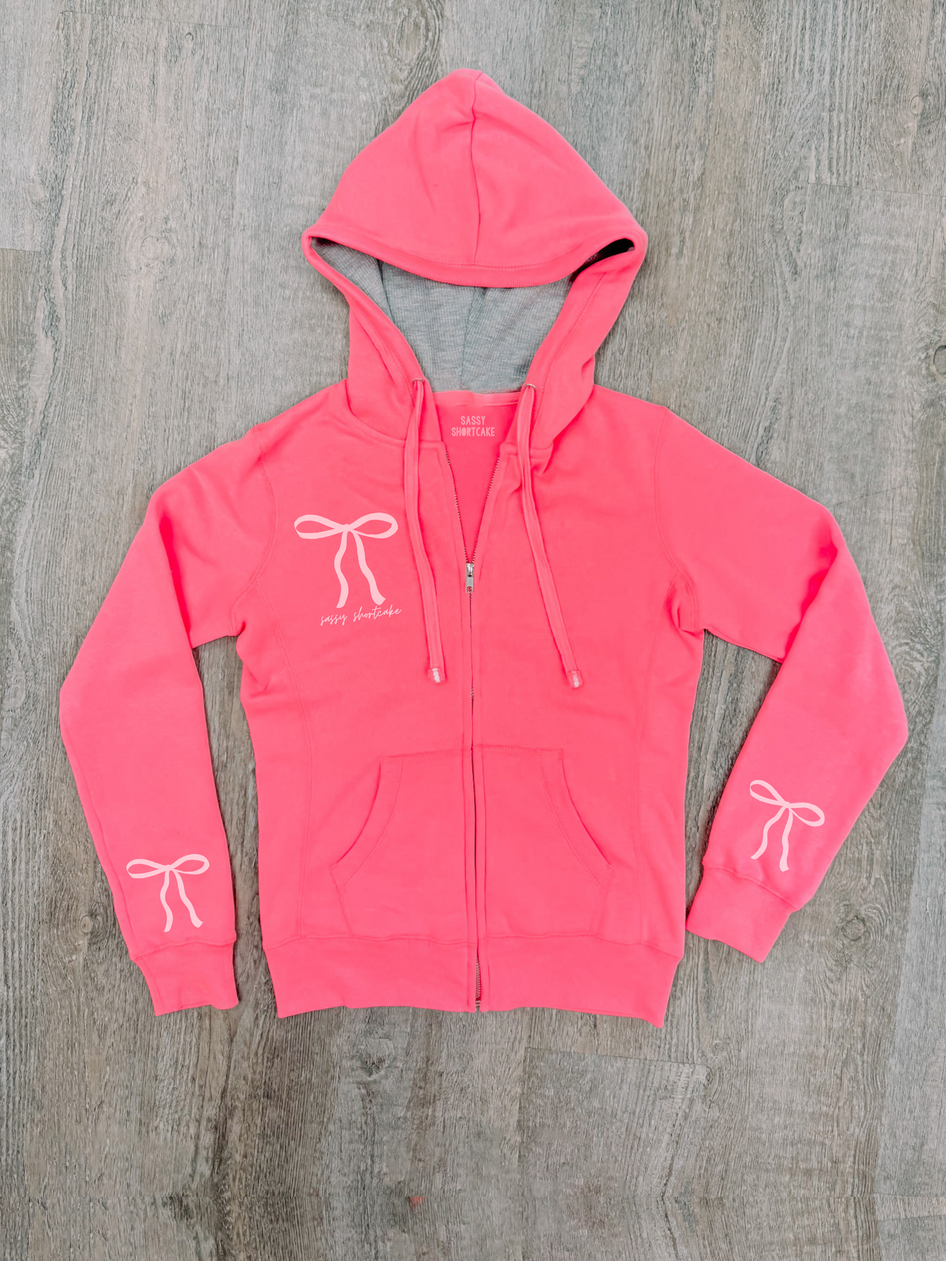 Neon pink zip-up hoodie with bow details, shown on wood background.
