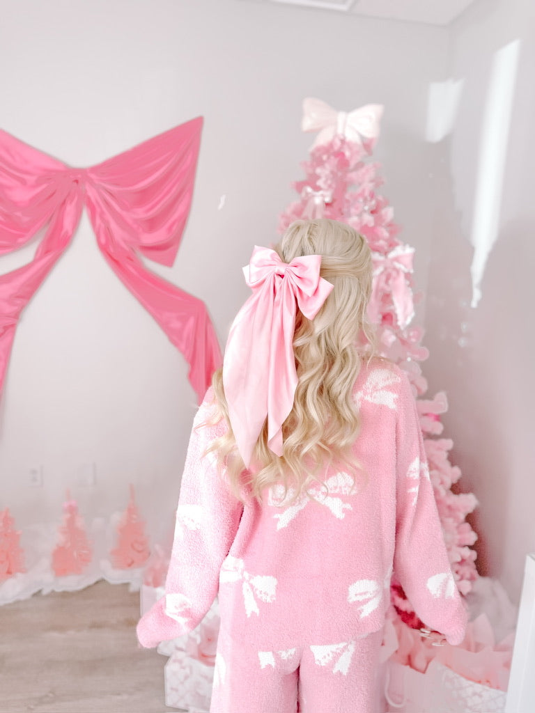 Pink fuzzy pajama set with bow pattern, worn with a pink satin ribbon bow in hair, near a pink Christmas tree.
