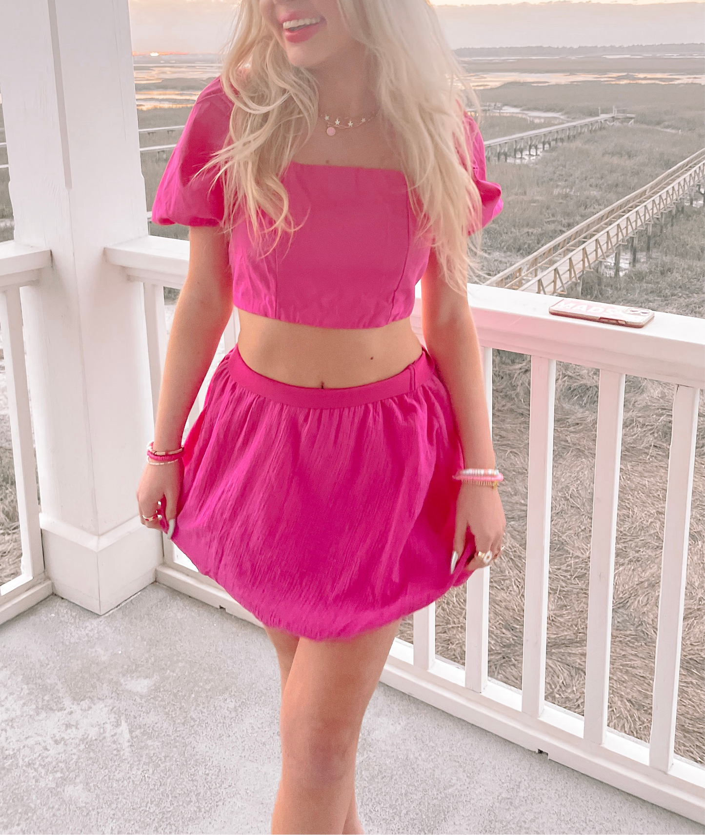 Hot pink mini skirt and matching crop top, worn outdoors on a porch overlooking a marshy landscape.
