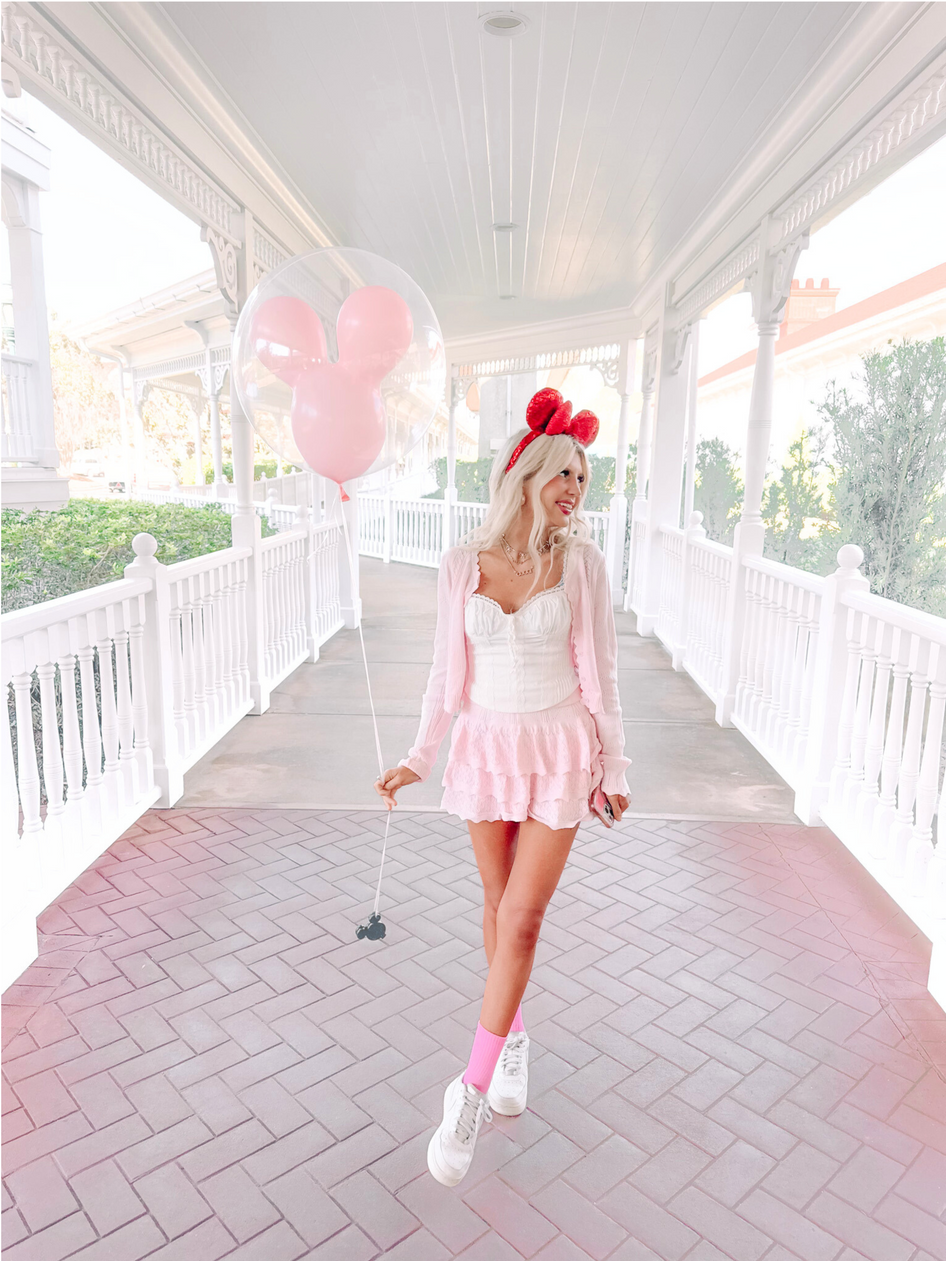 Woman in pink outfit on a white porch, holding Mickey Mouse balloons.
