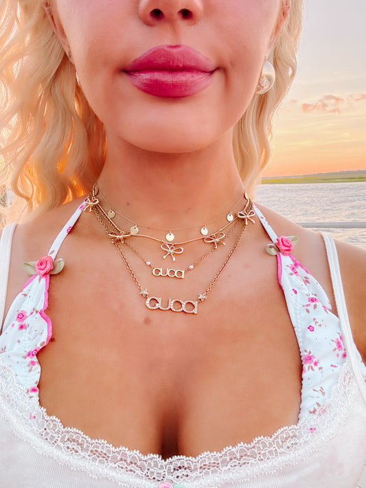 Gold layered necklaces with bow and lettering accents, worn against a floral top at sunset.
