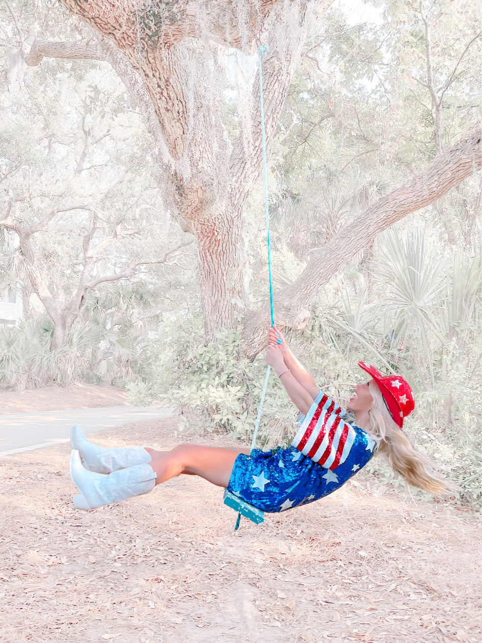 Woman in a patriotic sequin dress swinging from a tree outdoors.
