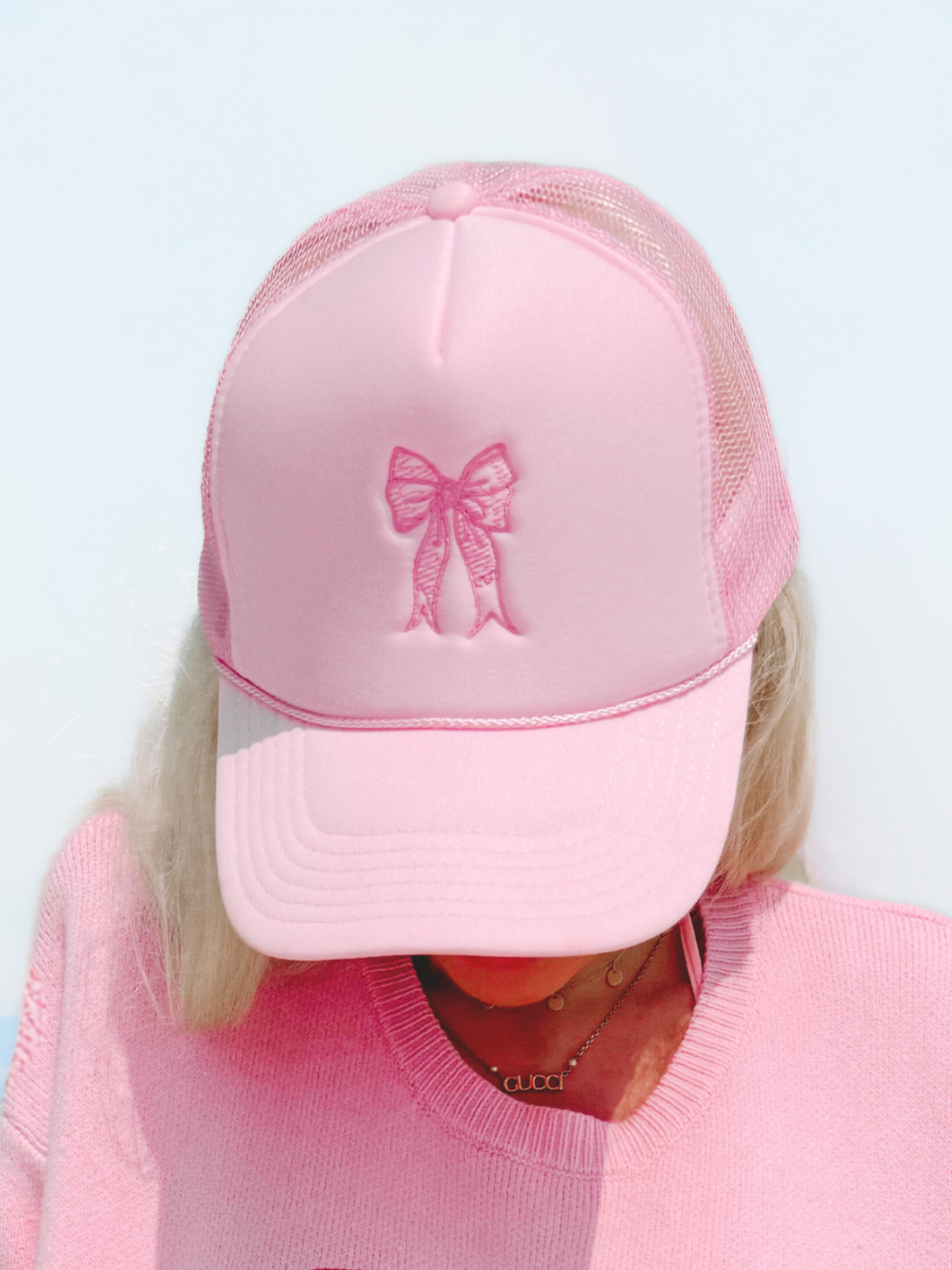 Pink trucker hat with embroidered bow, worn by blonde model in pink sweater.
