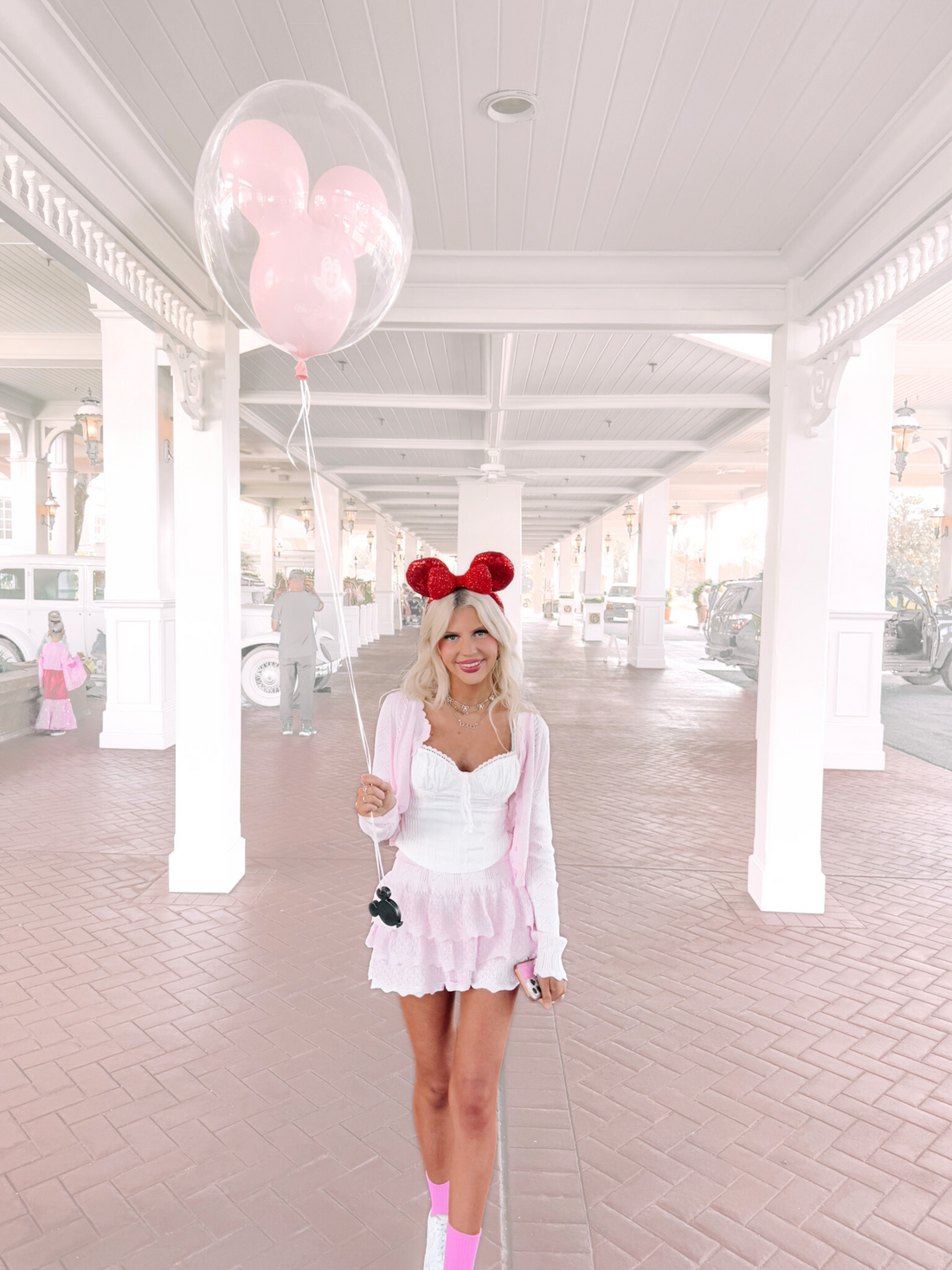 Woman in pink outfit and Minnie Mouse ears holding a pink Mickey Mouse balloon, walking under a white portico.

