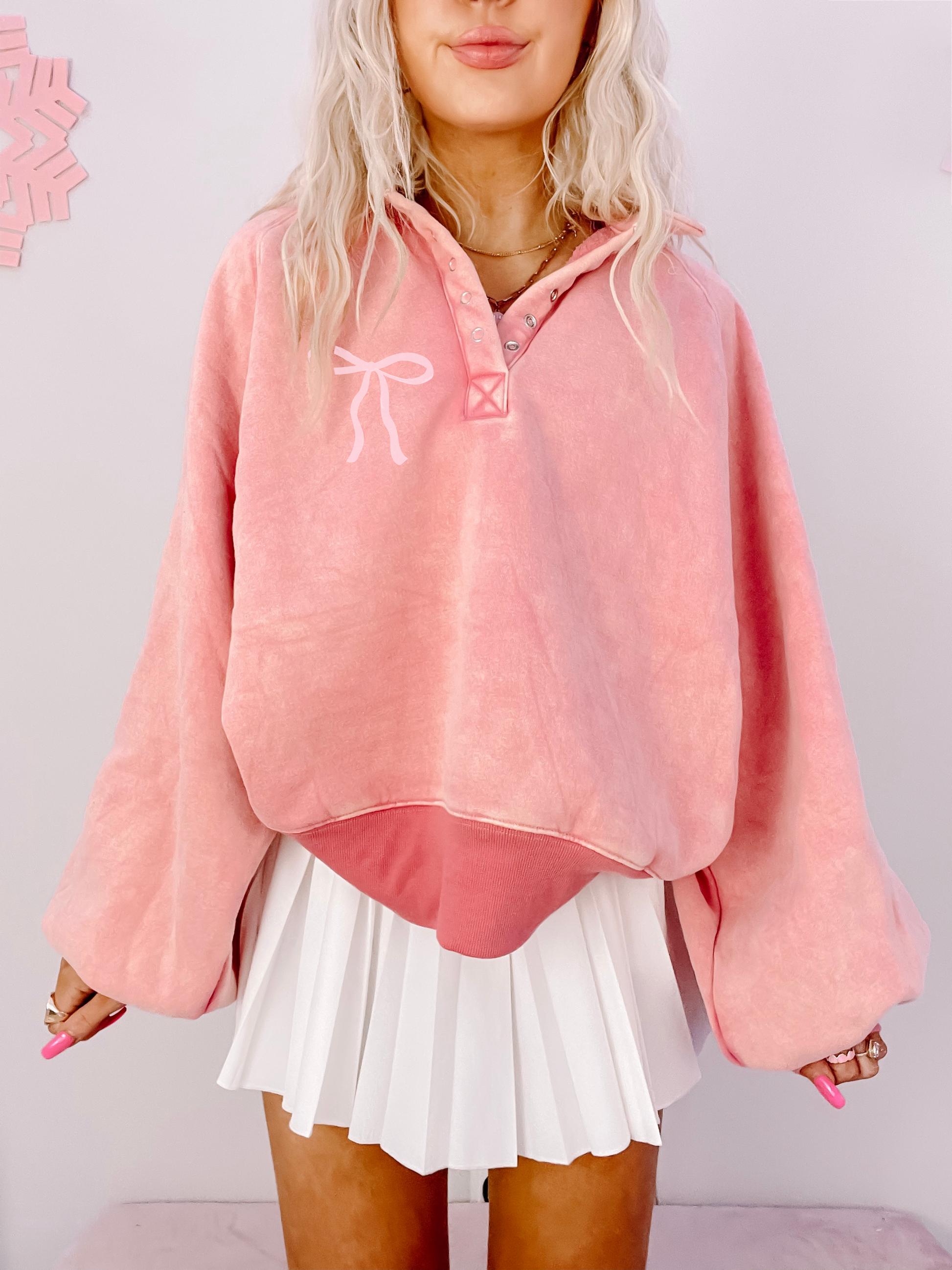 Pink pullover sweatshirt with a bow detail, styled with a white pleated skirt.
