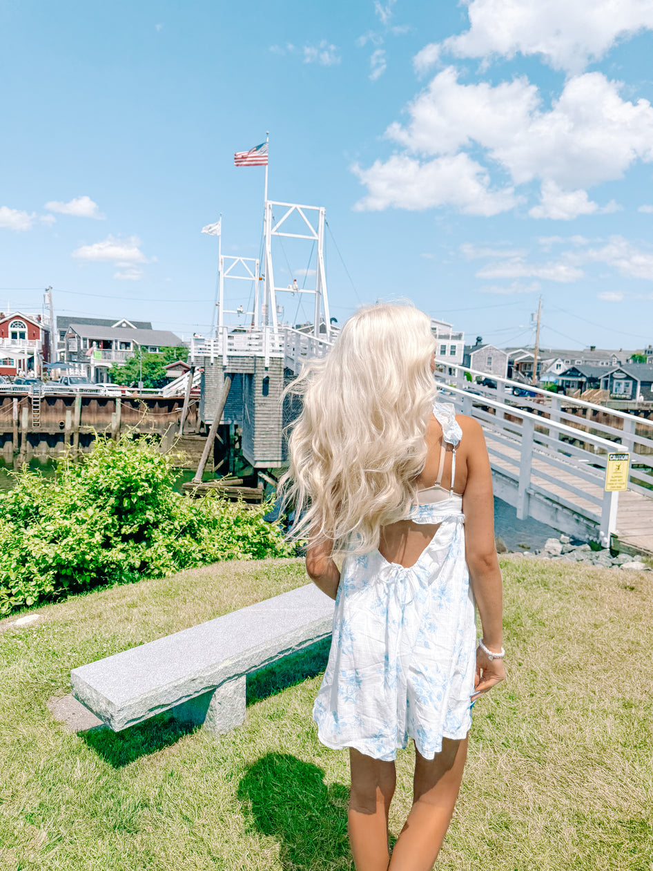 Woman in light blue floral dress, viewed from behind, near a waterfront with a white bridge and American flag.
