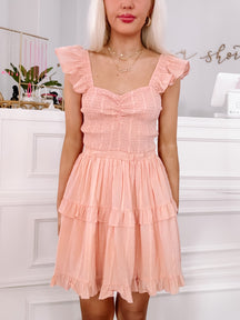 Peach mini dress with ruffled layers and smocked bodice, shown on a model in a boutique setting.
