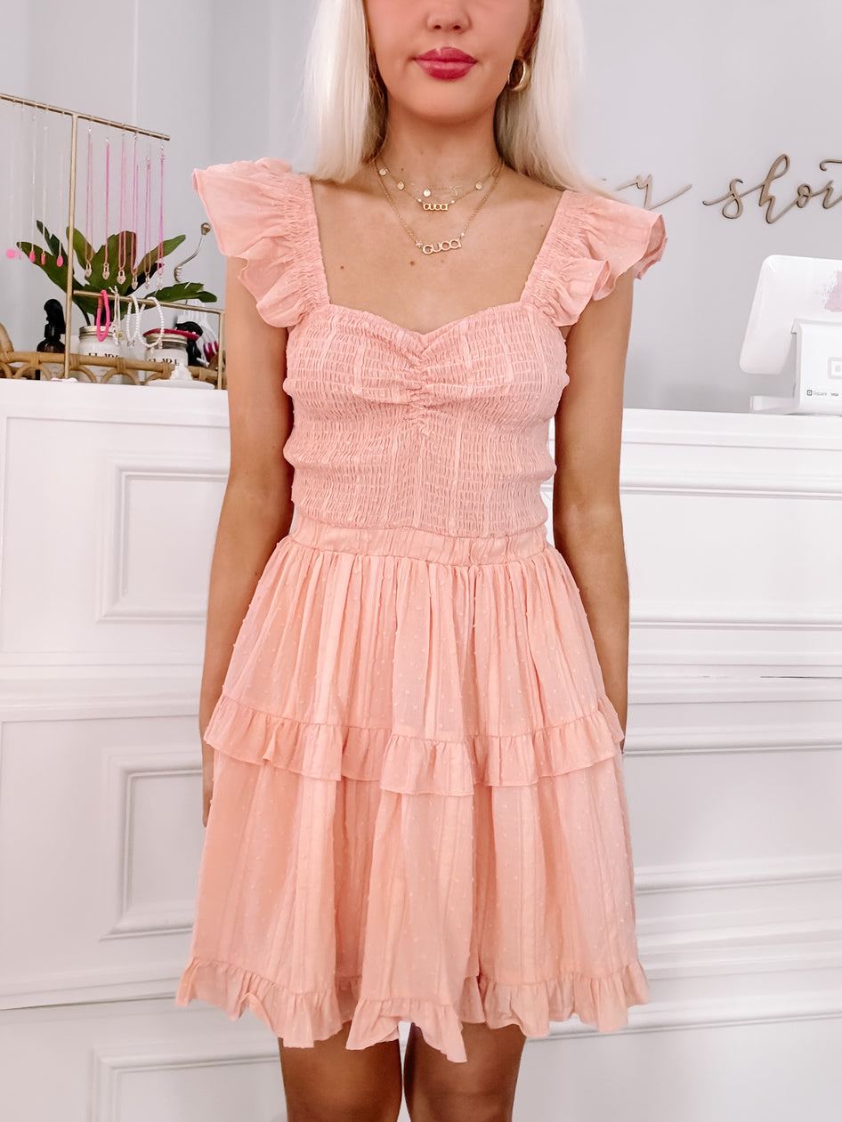 Peach mini dress with ruffled layers and smocked bodice, shown on a model in a boutique setting.
