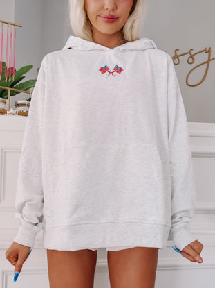 Model wearing light grey hoodie with embroidered American flag patch and cozy fit.