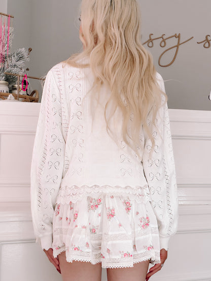 Bisous Bow Cardigan from Sassy Shortcake -Woman wearing a white lace outfit with floral details in a room setting.