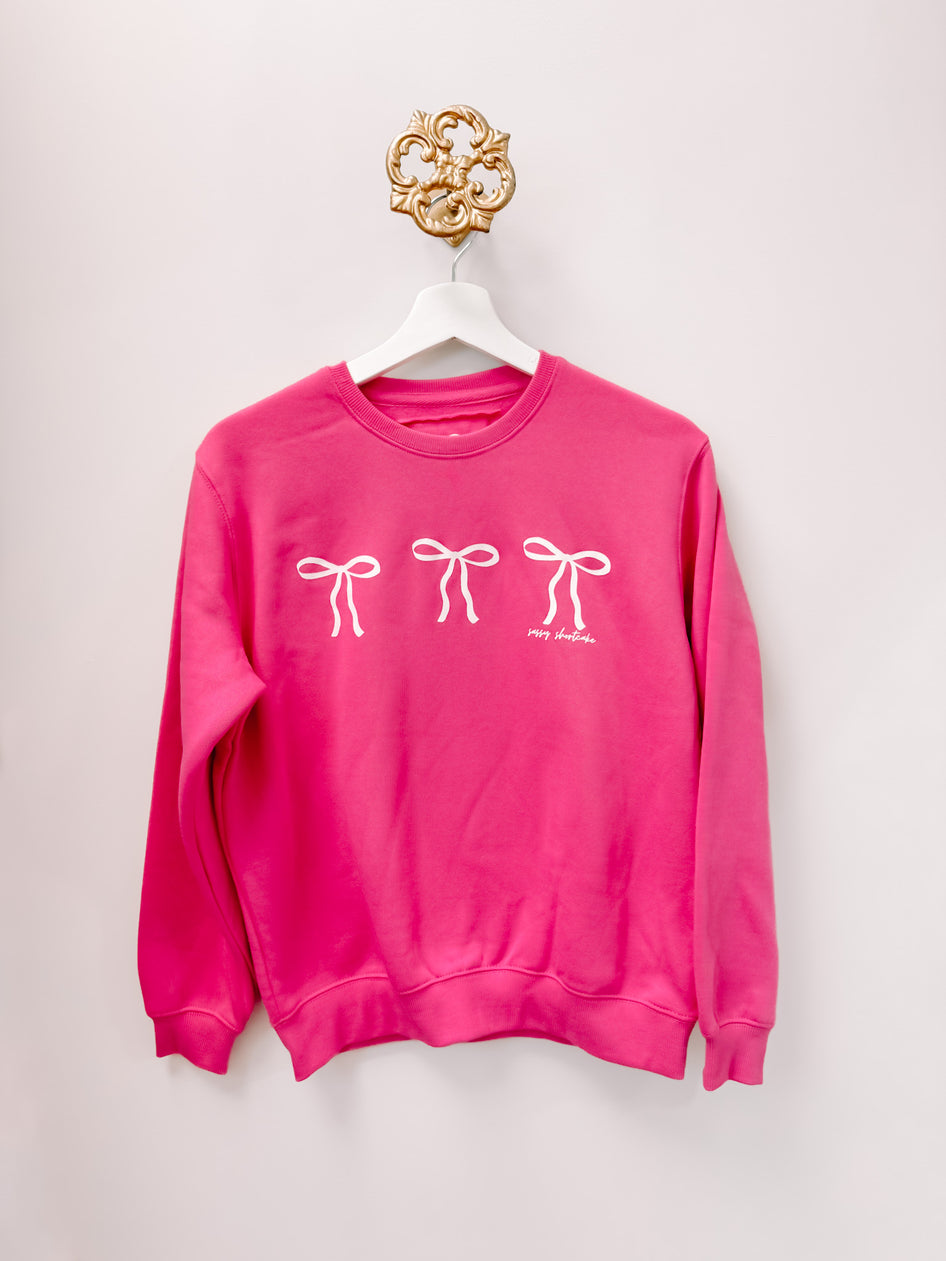Pink kids' crewneck sweatshirt with three white bows, hanging on a white hanger against a white wall.
