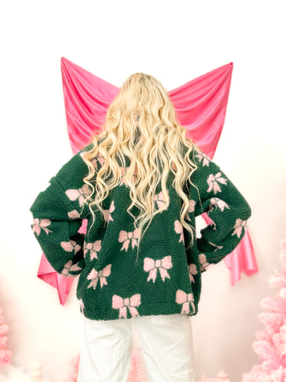 Woman modeling the cozy, textured hunter green sherpa bow pullover from Sassy Shortcake, featuring pink bows and functional pockets.