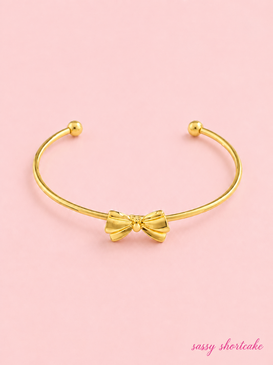 Golden Girl Bow Bracelet from Sassy Shortcake: Gold open cuff bracelet with a dainty bow detail on a light pink background