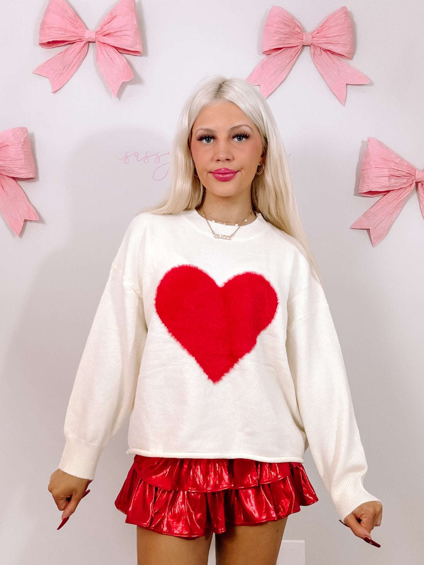 Cream sweater featuring a fluffy red heart, modeled against a backdrop of pink bows.
