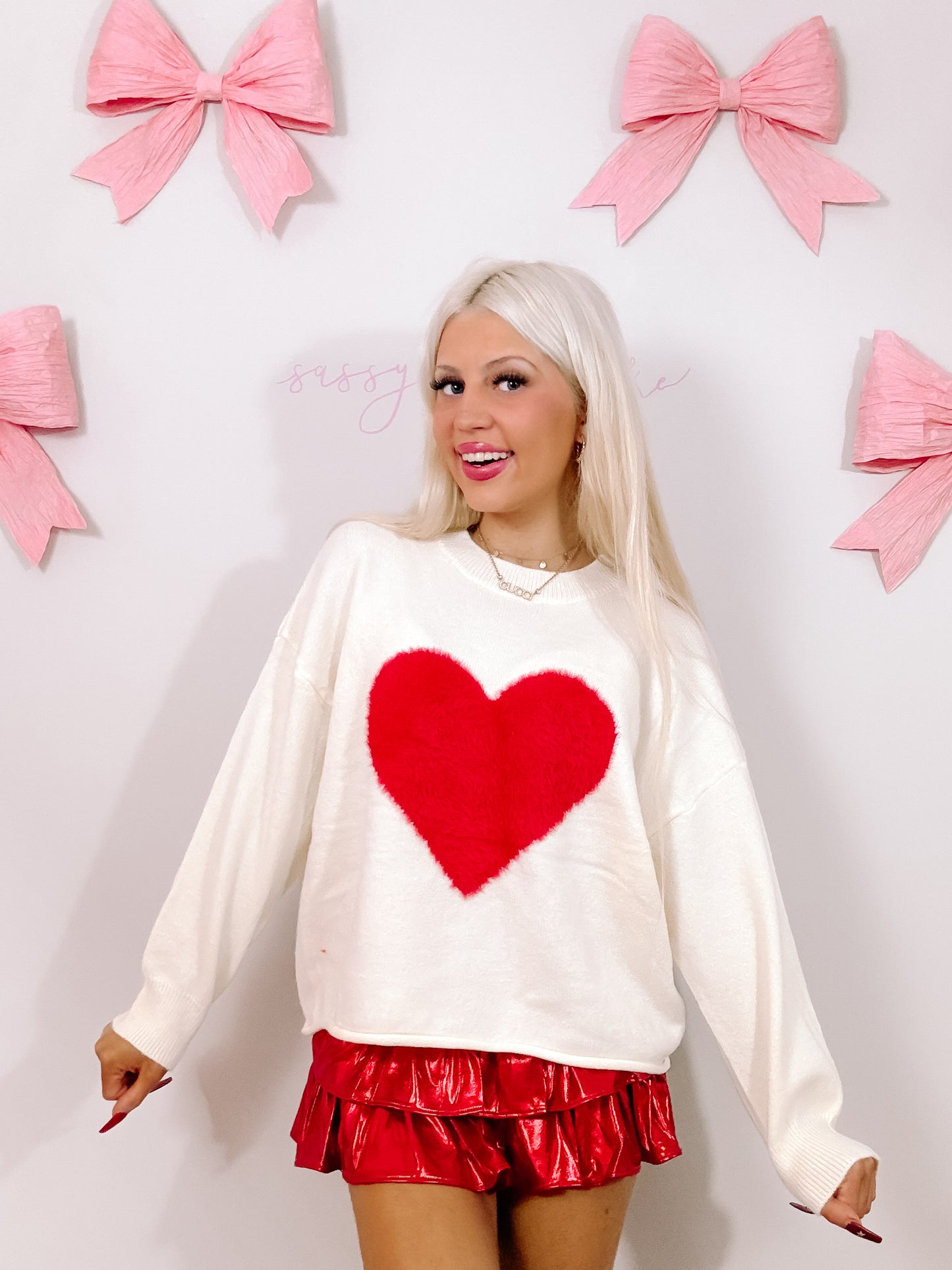 Cream oversized sweater featuring a fluffy red heart, modeled against a pink background.
