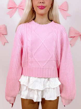 Pink cable-knit sweater with balloon sleeves, styled with a white ruffled skirt against a pastel background.
