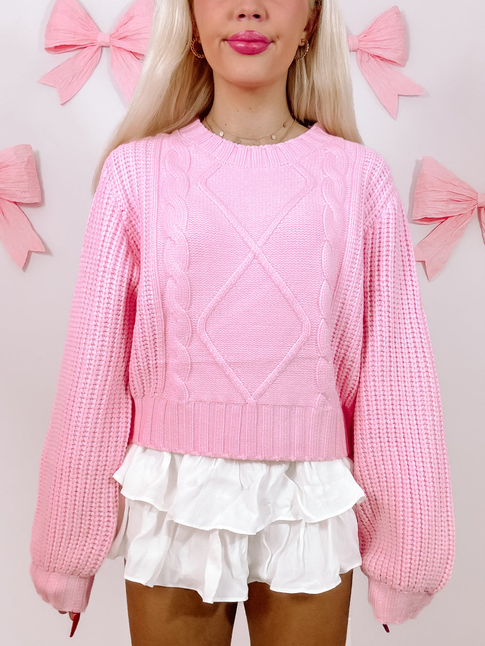 Pink cable-knit sweater with balloon sleeves, styled with a white ruffled skirt against a pastel background.
