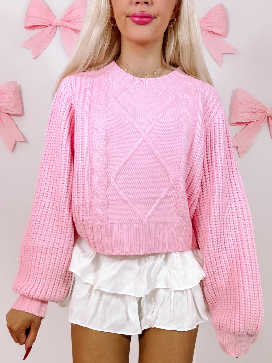 Pink cable-knit sweater with balloon sleeves, styled with a white ruffled skirt against a pastel background.
