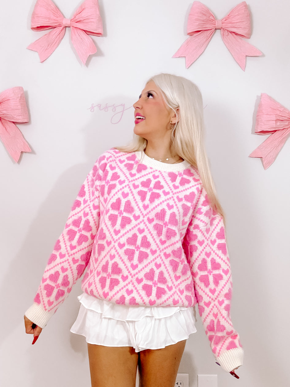 Pink fuzzy sweater with heart pattern, modeled against a wall with pink bows.
