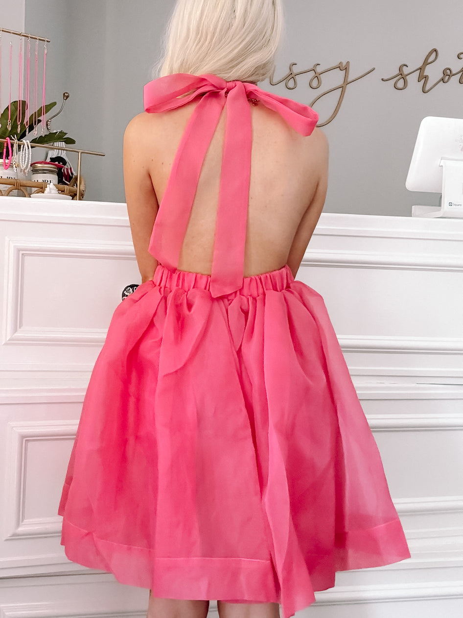 Hot pink halter mini dress with a bow detail, shown from the back in a boutique setting.
