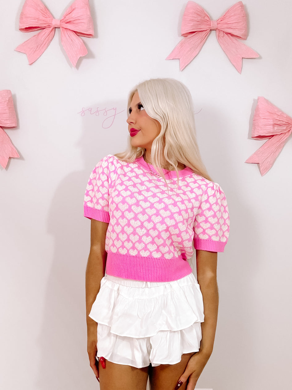 Model showcasing a pink heart-print top and white ruffled shorts against a backdrop of pink bows.
