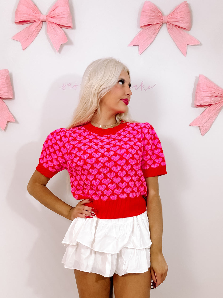 Red short-sleeve top with pink hearts, modeled against a white background with pink bows.
