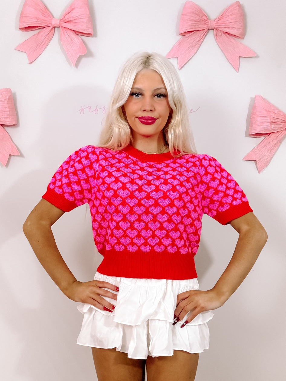 Red knit top adorned with pink hearts, short sleeves, modeled against a pink and white background.
