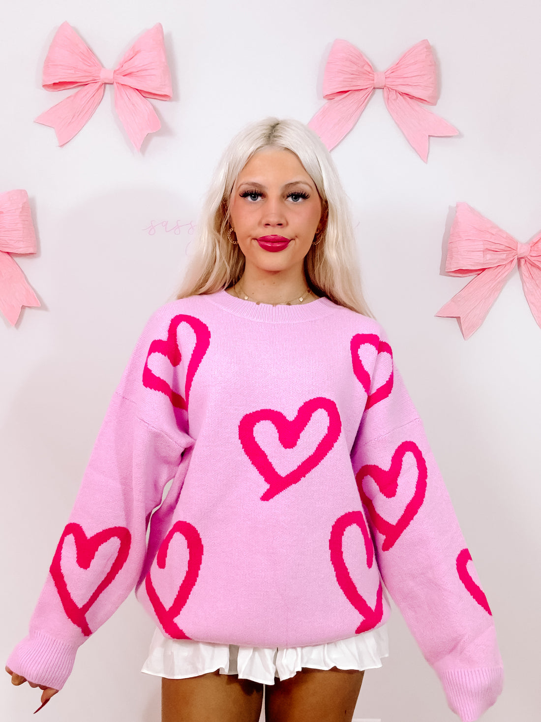Pink knit sweater adorned with bright pink heart patterns, styled against a backdrop of pastel pink bows.
