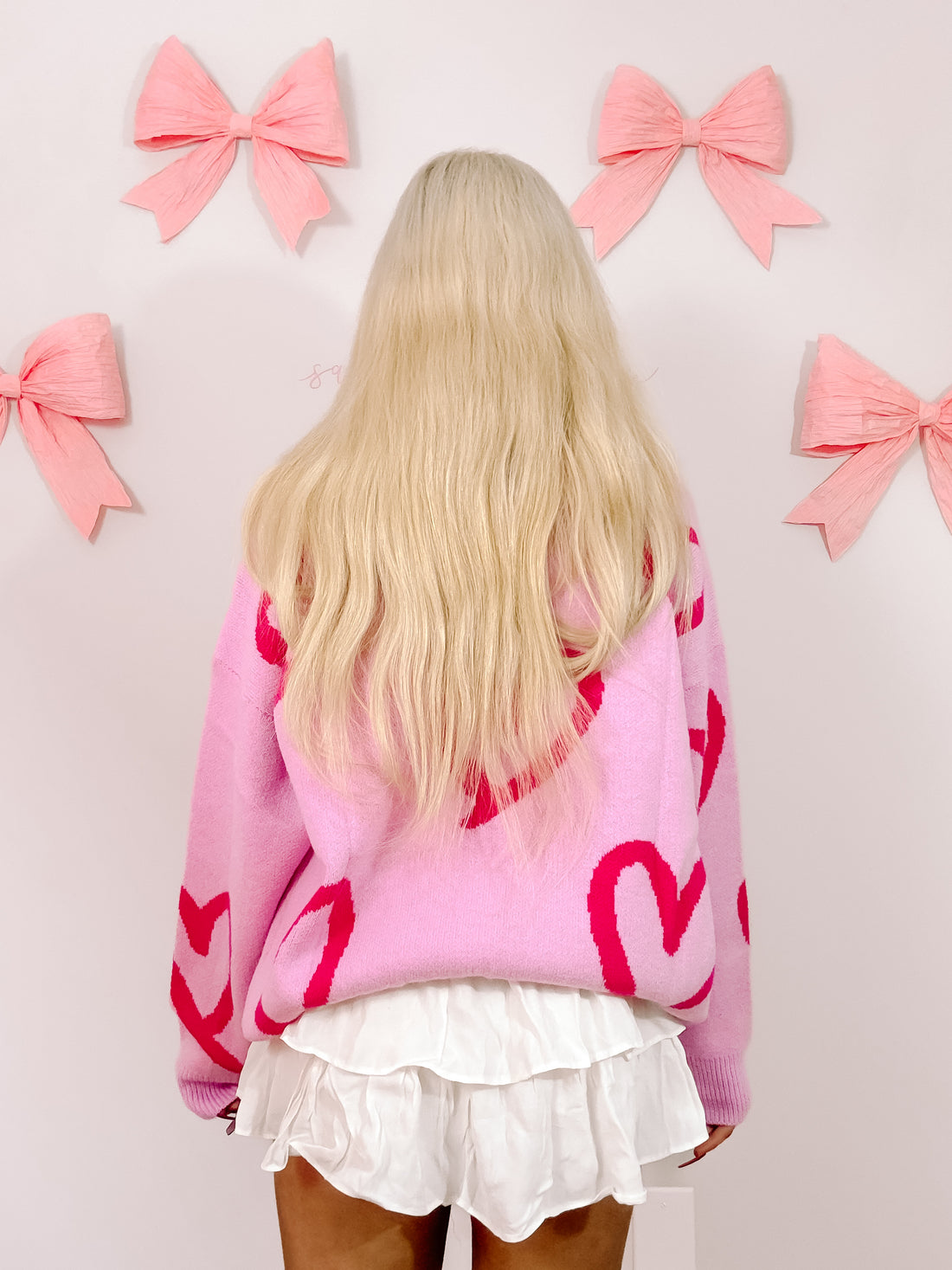 Pink knit sweater featuring bold red hearts, shown from the back against a wall adorned with pink bows.
