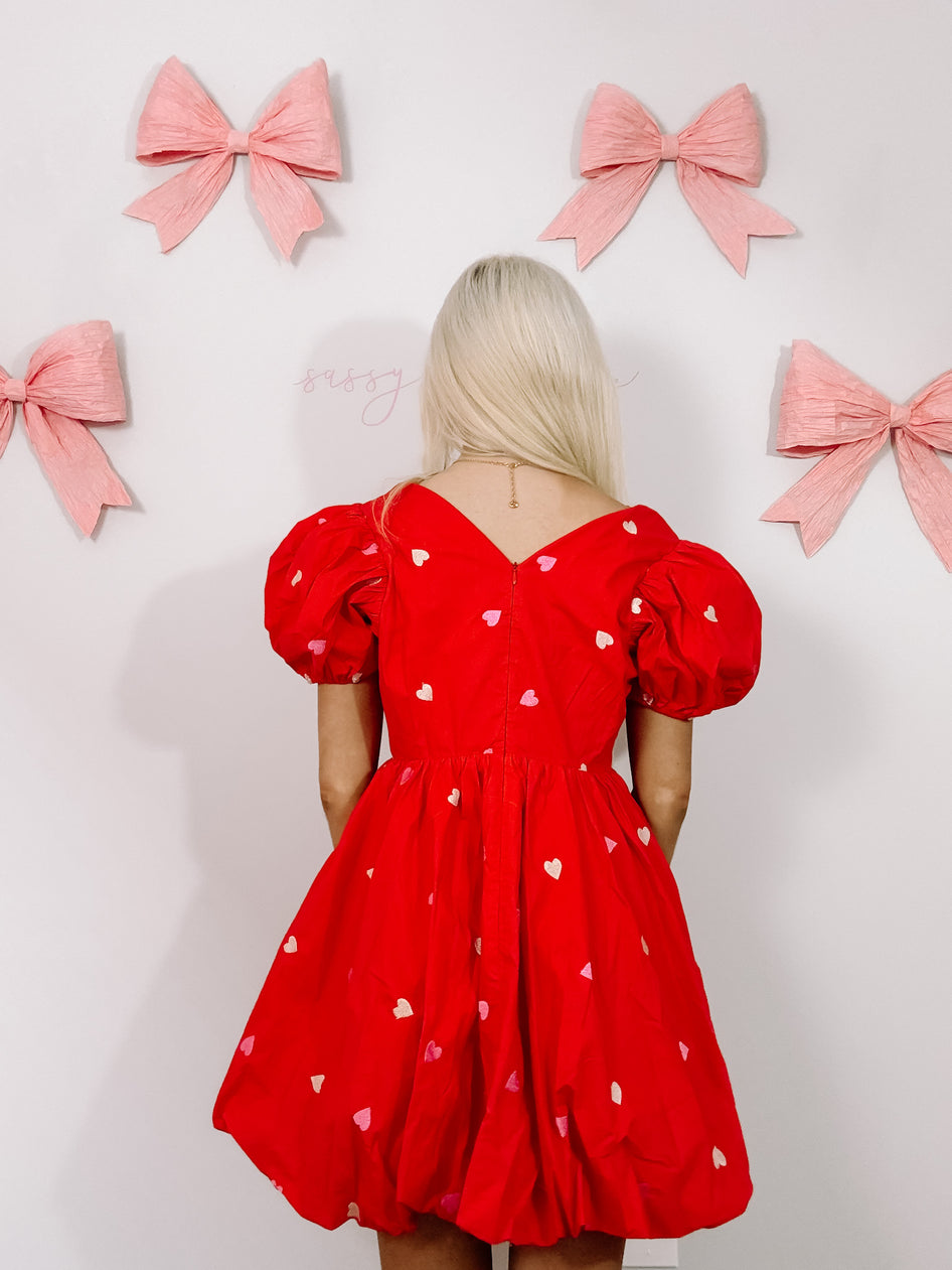 Red heart-print mini dress with puff sleeves, shown from the back against a wall decorated with pink bows.
