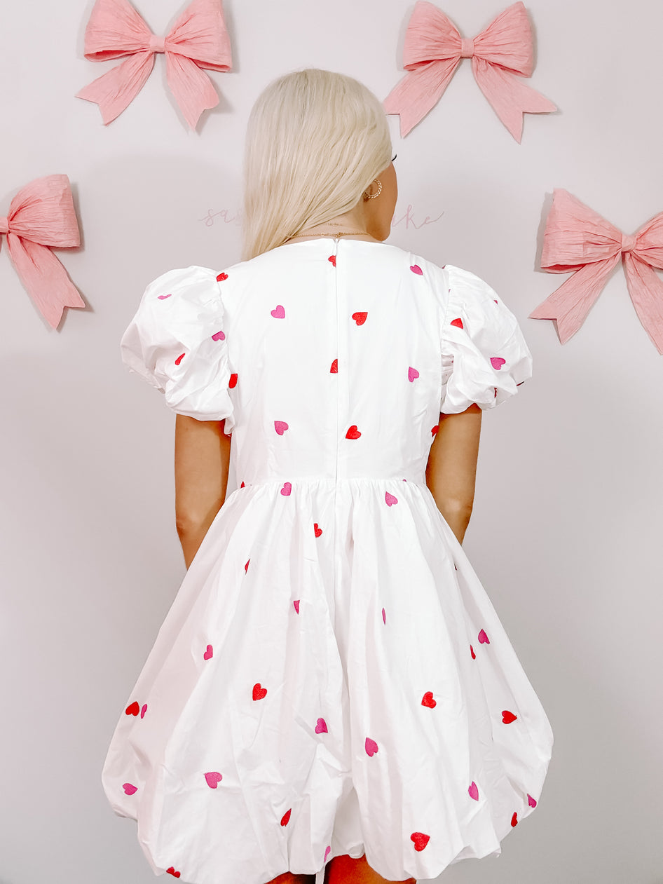 White puff-sleeve mini dress adorned with scattered pink and red hearts, shown from the back against a light wall.
