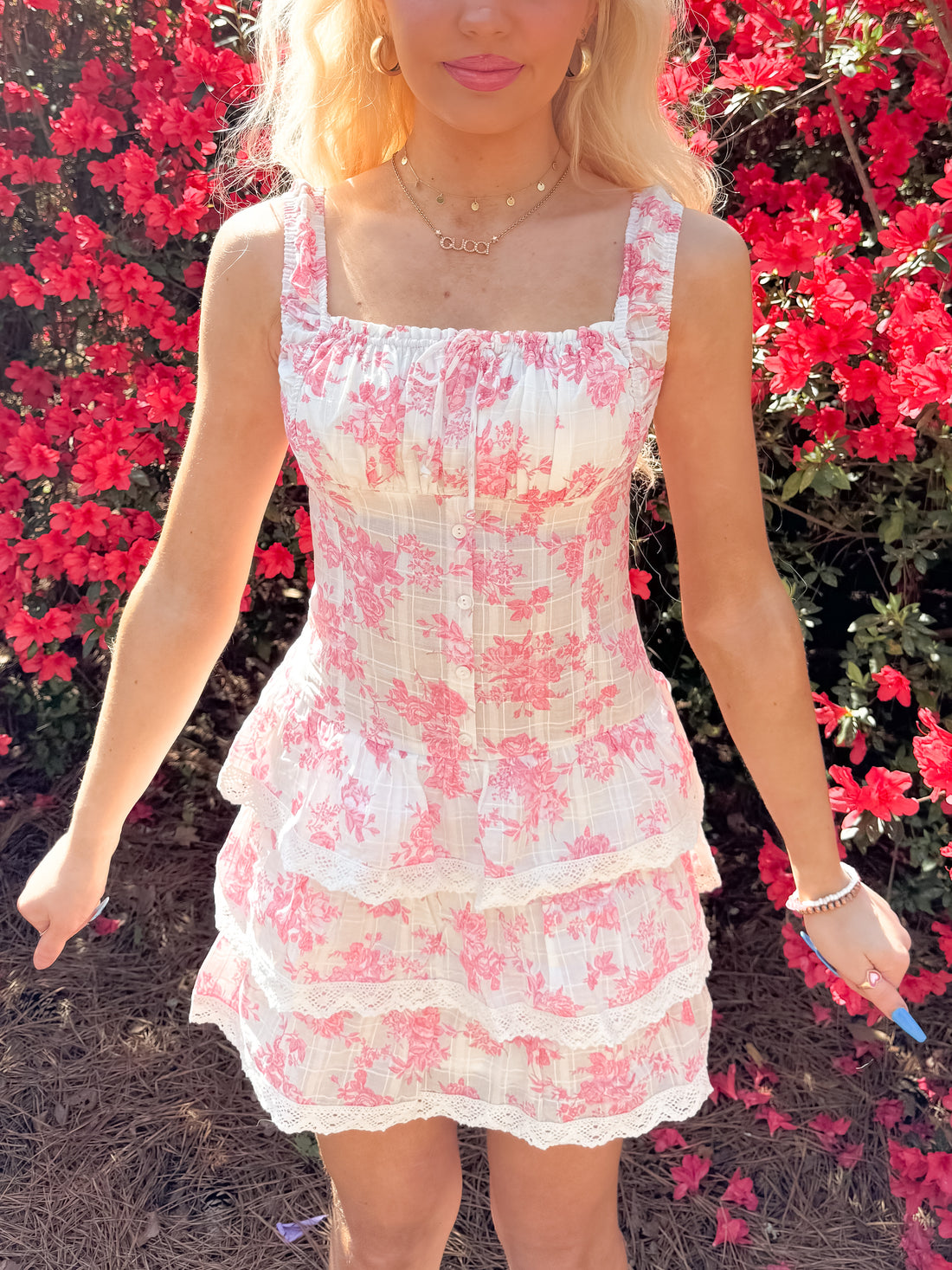 Pink floral mini dress with tiered ruffle skirt, worn outdoors amongst vibrant red flowers.
